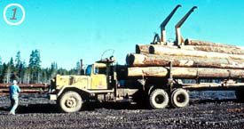 The carrier truck approaches from the far side and places carrier mechanism over load and secures it for the approaching truck drivers. Note: The load binders are still secured in place.